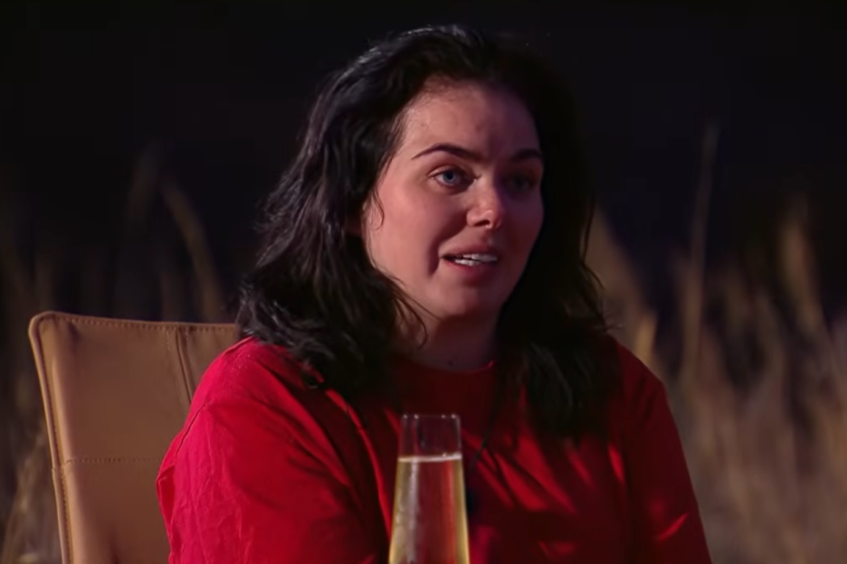 A woman with dark hair and a red top, sitting in a chair with a glass of champagne in front of her.