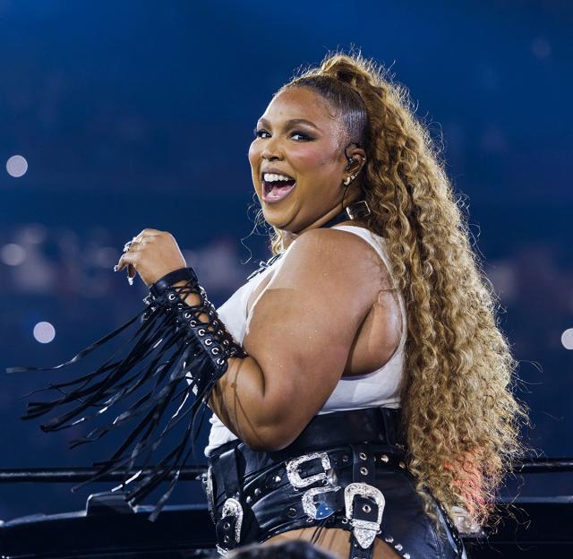 Lizzo performs on stage, wearing a white top and black leather shorts with buckles, and a matching arm cuff.