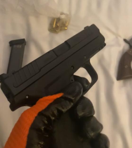 A person wearing a black and orange glove holding a black handgun, with a detached magazine and loose bullets in the background.