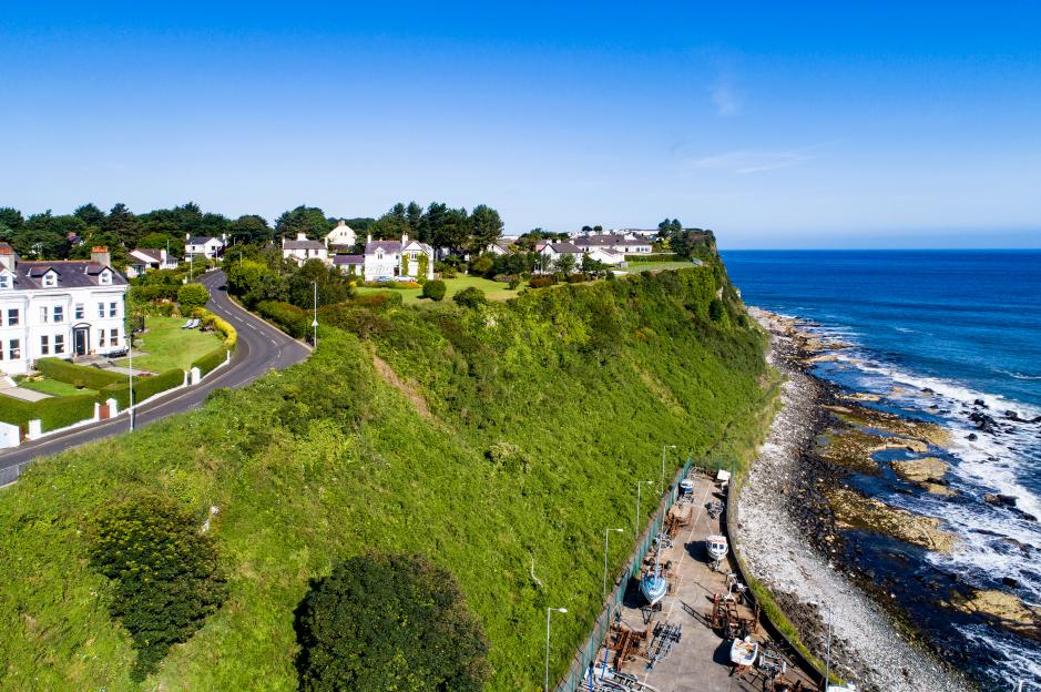 Aerial view of the Atlantic coast with a steep cliff, Ballycastle marina, and Causeway Coastal Road in Northern Ireland.