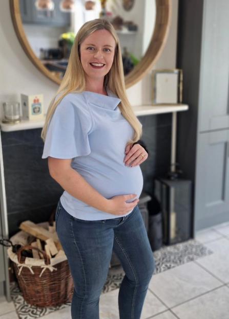 A pregnant woman in a light blue top and jeans smiles while holding her baby bump.