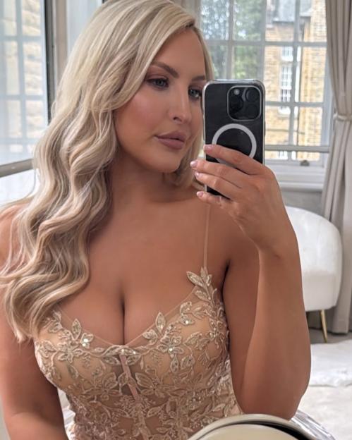 A blonde woman takes a mirror selfie, showing off a low-cut beige dress with intricate gold and silver embroidery.