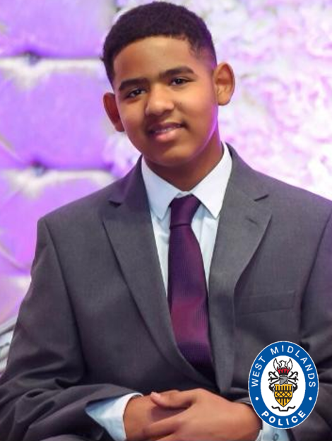 A young man in a gray suit and purple tie with a West Midlands Police emblem in the bottom right corner.