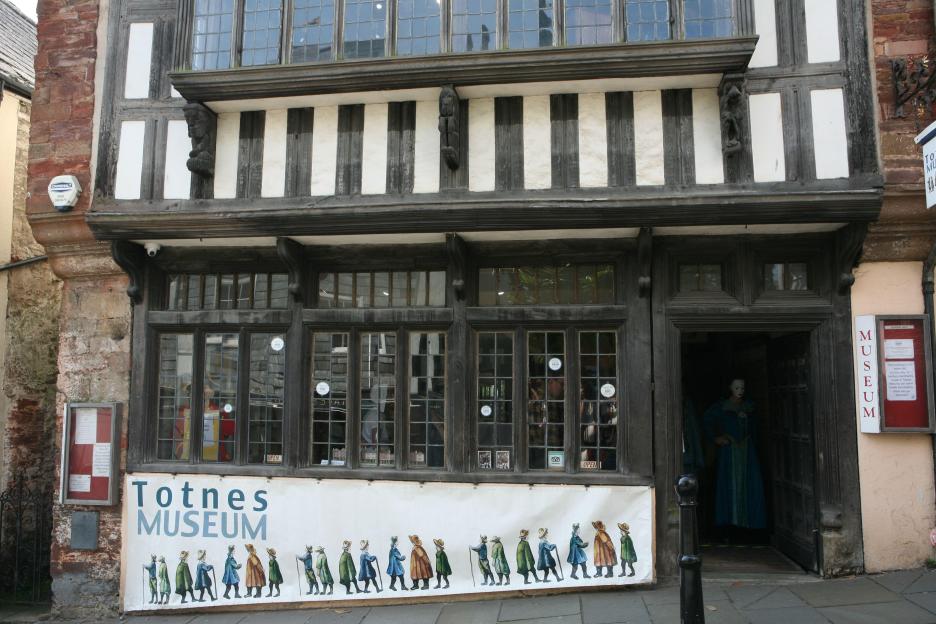 Exterior of Totnes Museum, a historic timber-framed building in England, with a banner featuring illustrations of people in historical clothing.