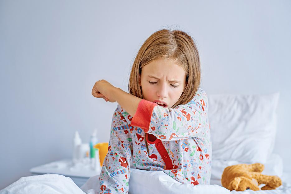 A young girl with long brown hair, wearing pajamas, coughs into her elbow while sitting in bed.