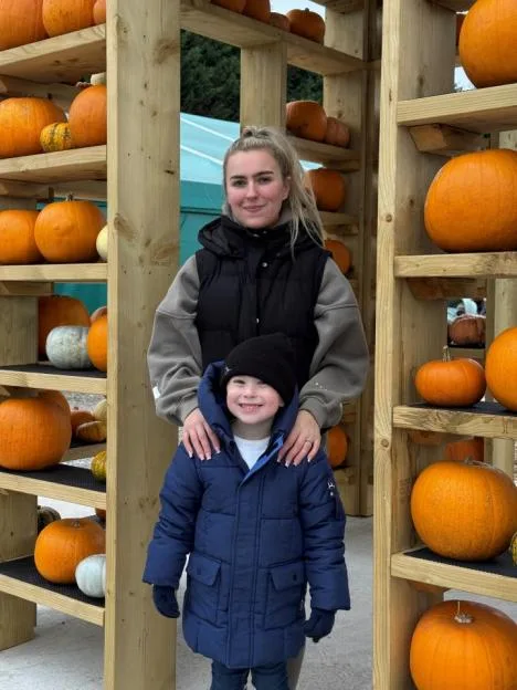 Jess Hulley-Jones with her son Theodore standing among wooden shelves filled with pumpkins.