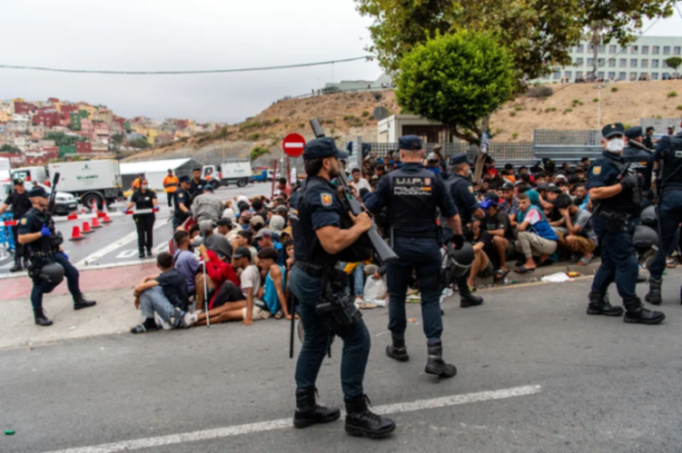 Armed police officers stand guard over a large group of people sitting on the ground.