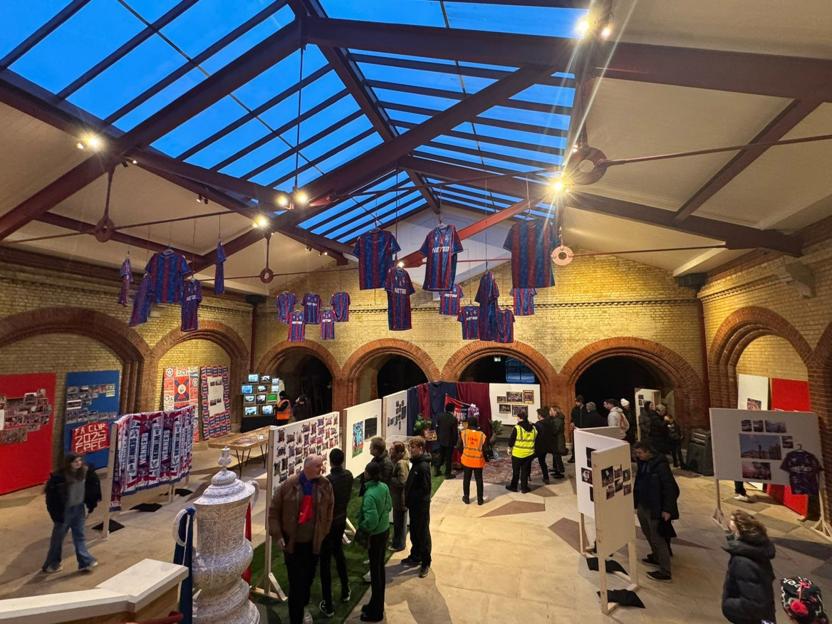 A Crystal Palace football club exhibition celebrating their FA Cup victory, with jerseys hanging from the ceiling, memorabilia on display, and fans viewing the exhibits.
