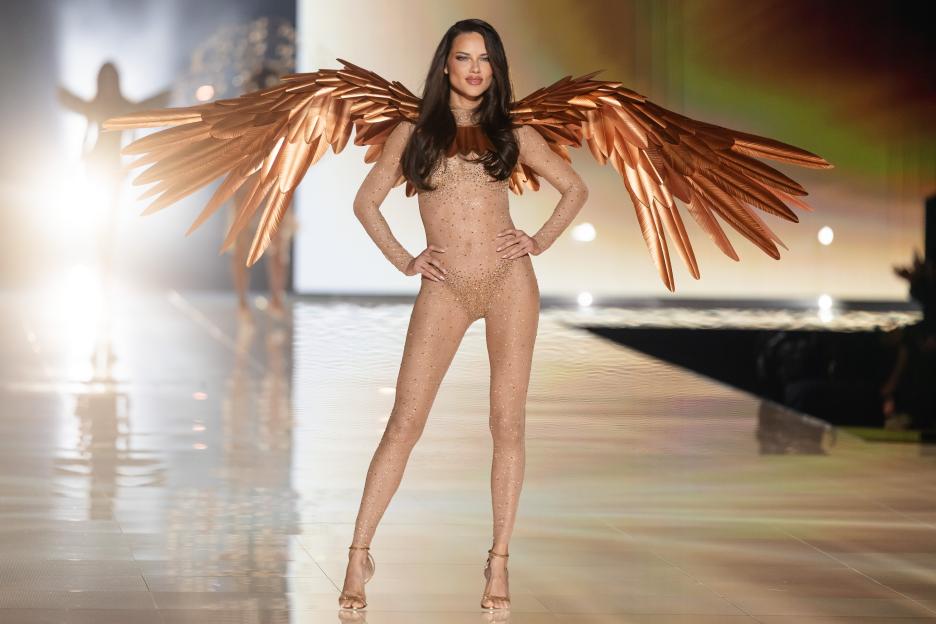 Adriana Lima walking the runway in a sheer, sparkling bodysuit and large, metallic copper wings.