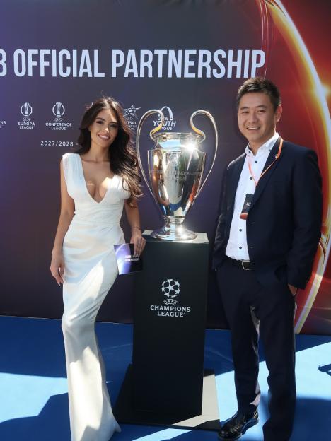 A man and woman standing next to the UEFA Champions League trophy with a "Partnership" banner in the background.