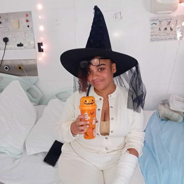 Ria Miswell, wearing a witch hat and an arm cast, holding a Halloween cup in a hospital bed.