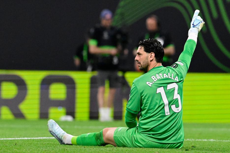 Soccer player in a green jersey and white gloves with arms outstretched on a field.