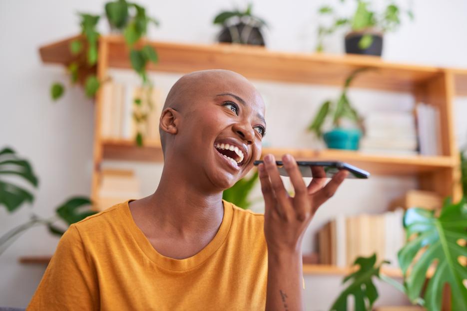 A young woman sends a voicenote the living room with a bookshelf of plants