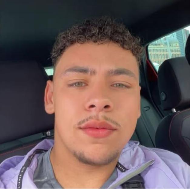 A young man with light green eyes, light brown curly hair, and a mustache and goatee, wearing a purple jacket over a gray shirt, in a car.