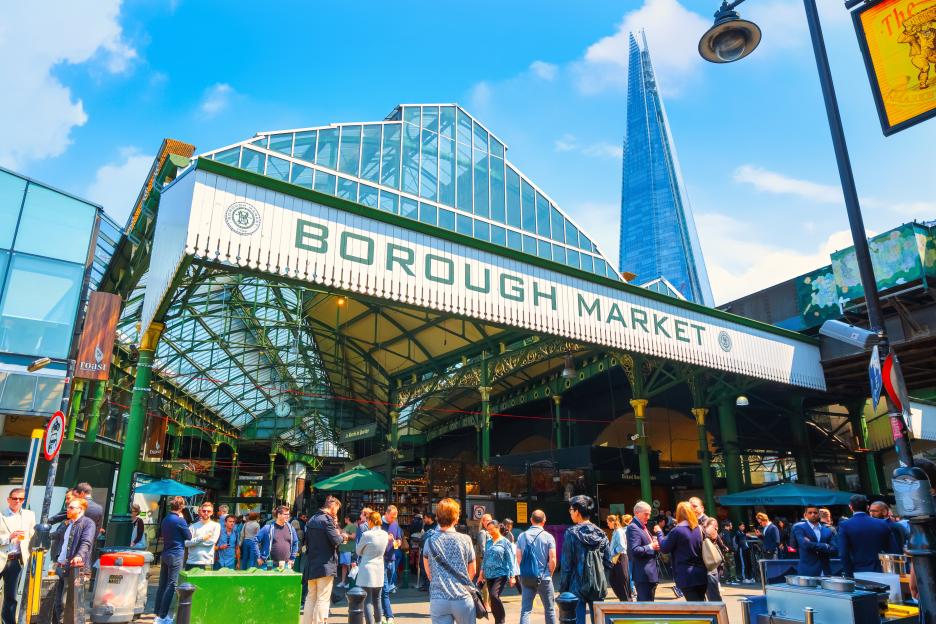 People gather under the "Borough Market" sign in London, with The Shard skyscraper visible in the background.