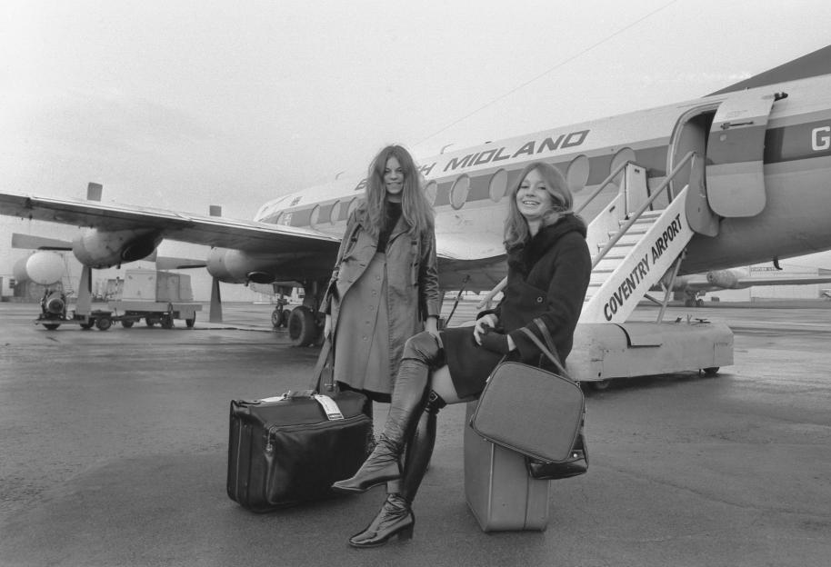 Two young women with luggage on an airport tarmac, with an airplane in the background.