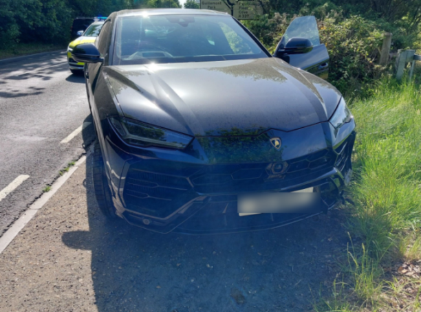 A black Lamborghini Urus with an open door parked on the side of a road, next to a police car.