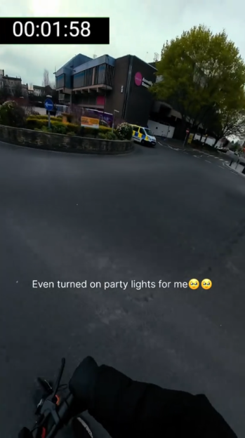 A person on an e-scooter approaches a roundabout with a police van with active lights on the far side.