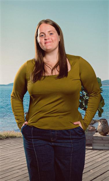 A woman wearing a mustard yellow top and dark blue jeans, standing on a wooden deck by the sea.