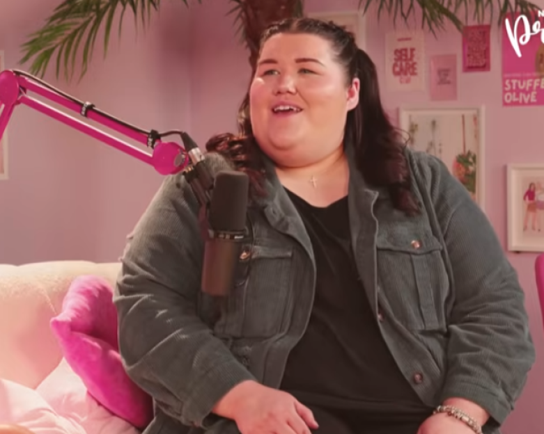 A woman with dark hair laughs while seated on a pink couch with a microphone in front of her.