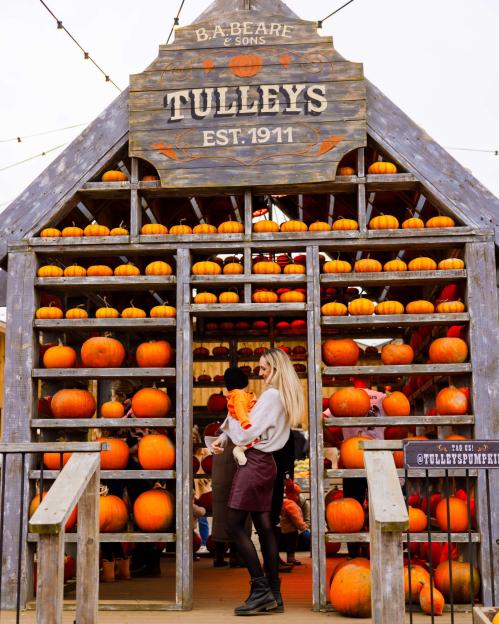 A blonde woman holding a baby stands in front of a pumpkin stand with "Tulleys Est. 1911" written on a sign.