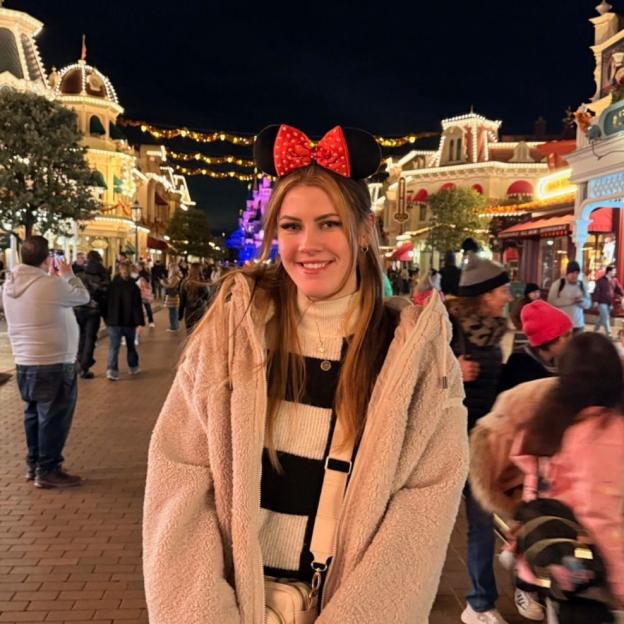 A woman wearing Minnie Mouse ears smiles at night in a busy theme park.