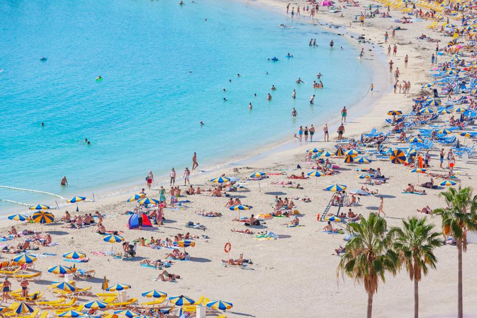 Amadores beach in Gran Canaria with many people swimming, sunbathing under umbrellas, and palm trees.