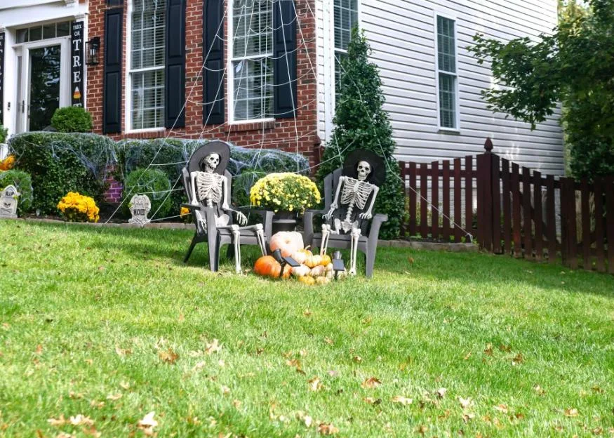 Two skeletons sitting in chairs on a front lawn decorated for Halloween with pumpkins and spider webs.