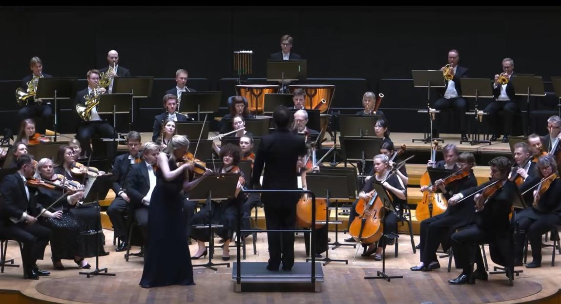An orchestra performing on stage, with a conductor on a podium in the foreground and a female soloist playing violin in the center.