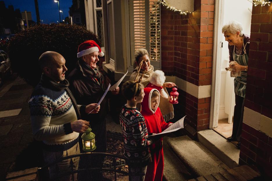 A multi-generational family singing Christmas carols at a front door, as a senior woman opens it smiling.