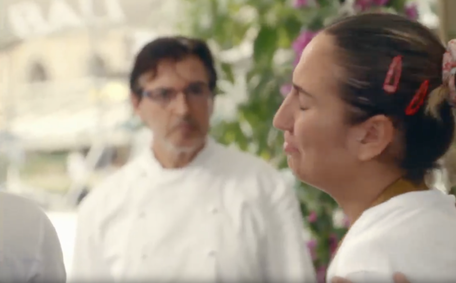 A woman crying with her eyes closed, while a man in a chef's uniform looks on in the background.