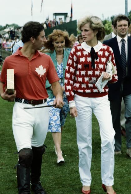 Princess Diana in a red and white sheep sweater, Prince Charles in a red polo shirt, and Sarah Ferguson in a floral dress walk on grass at a polo meeting.