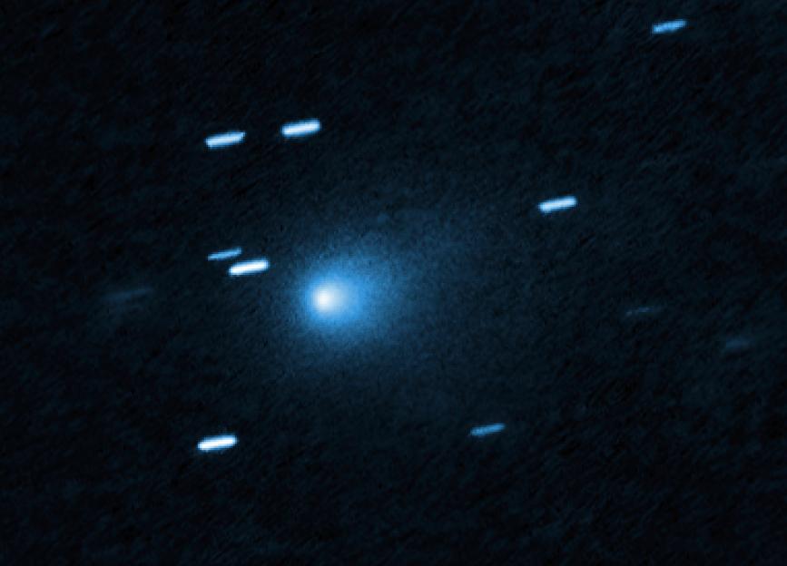 Hubble image of interstellar comet 3I/ATLAS, a bright blue-white point of light, surrounded by smaller, streaked objects, against a dark blue background.