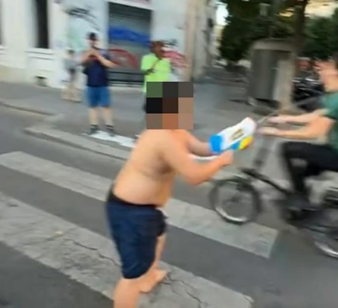 Teenager with a pixelated face, barefoot and shirtless, in dark shorts, holding a water pistol on a crosswalk as a person on a bicycle passes by.
