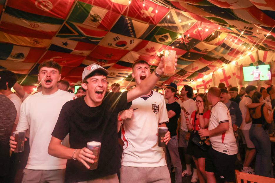 England supporters cheer while watching a football match on television in a pub decorated with flags.