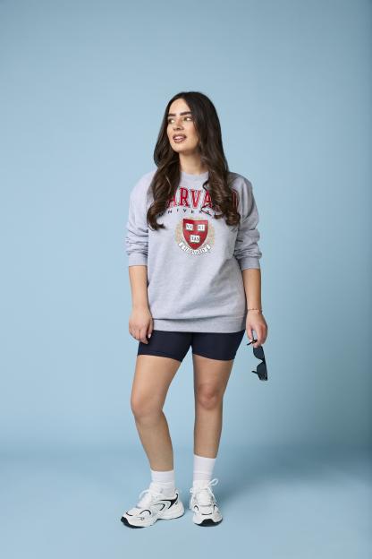 Emily Regan modeling a gray Harvard sweatshirt, black bike shorts, white socks, and white sneakers, holding sunglasses.