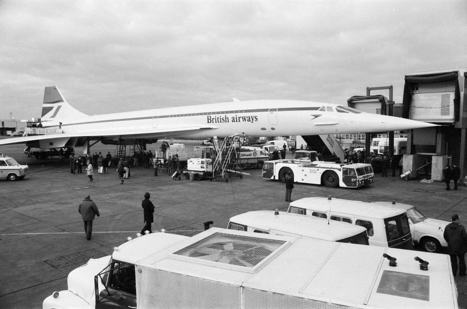 A black and white image of a British Airways Concorde aircraft at an airport terminal with people and service vehicles on the tarmac.