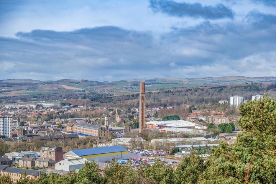 Panoramic view of the city of Dundee, including buildings, industrial areas, a large chimney, and distant hills.