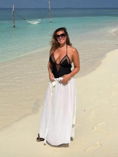 A woman in a black swimsuit and white skirt standing on a beach with clear blue water and a hammock in the background.
