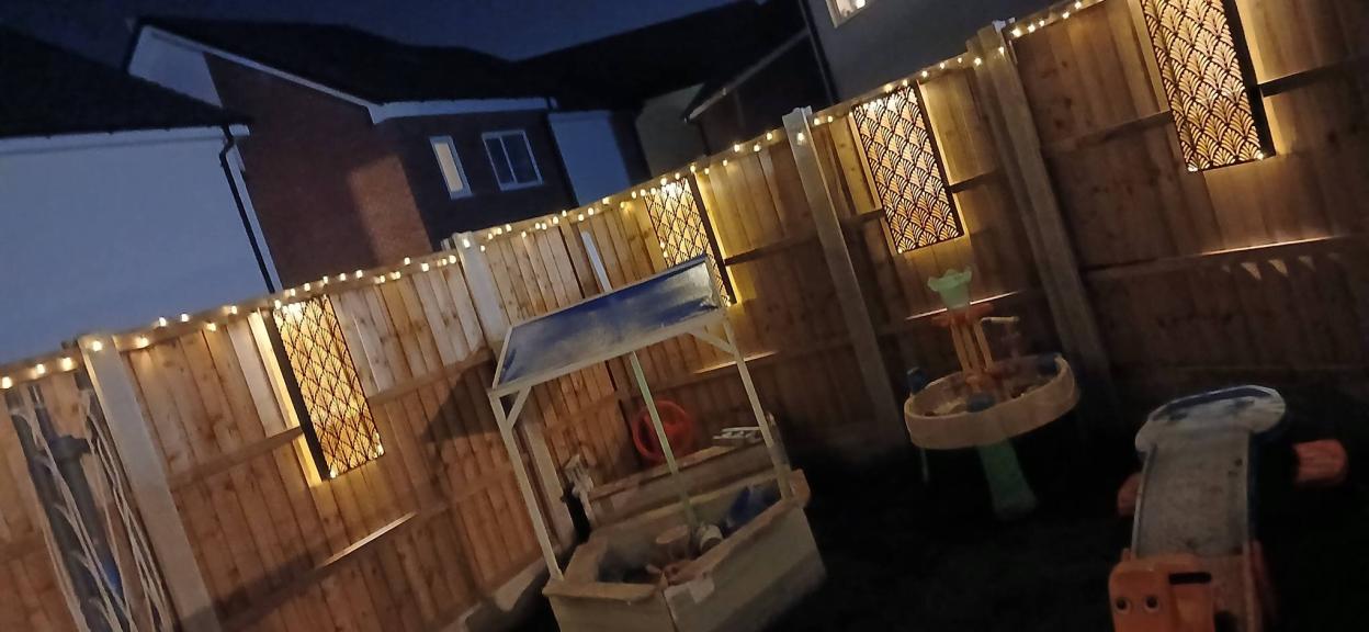 A backyard fence illuminated with string lights and patterned wall decorations.