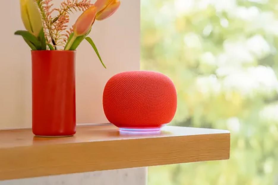 A red Google Home smart speaker sits on a wooden shelf next to a red vase with flowers.