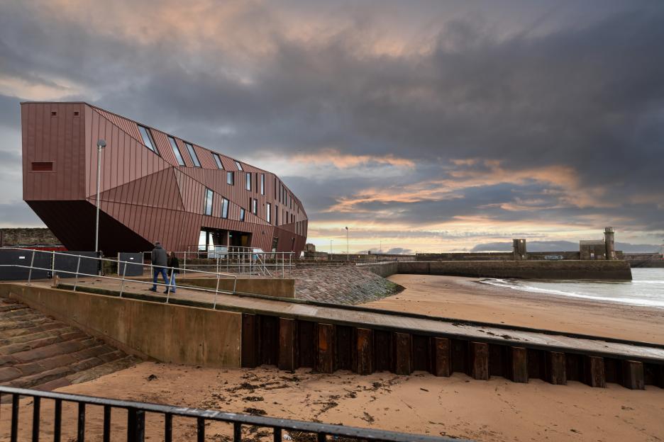A modern, angular brown building with many windows by a sandy beach, with a stone pier and cloudy sky.
