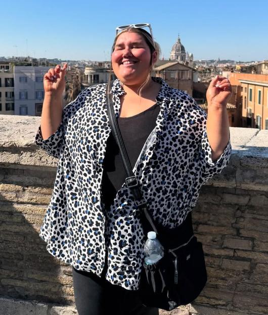 Becky Jones stands on a stone wall overlooking a city, wearing a white and black spotted shirt and black top, smiling with both hands raised.