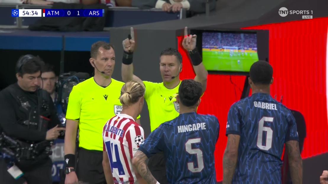 A referee points to a screen showing a football match replay, watched by players and another official.