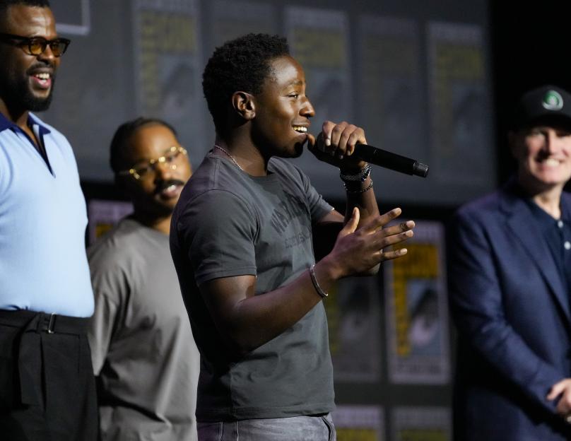 David Jonsson speaks into a microphone at Comic-Con, flanked by other men on stage.