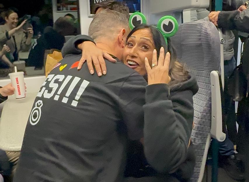 A woman with a frog-eye headband, an engagement ring, and a surprised expression embraces a man in a "YES!!!" shirt on a train.