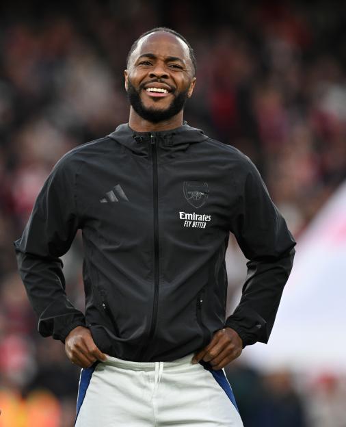 Raheem Sterling smiling while wearing an Arsenal jacket and white shorts at Emirates Stadium.