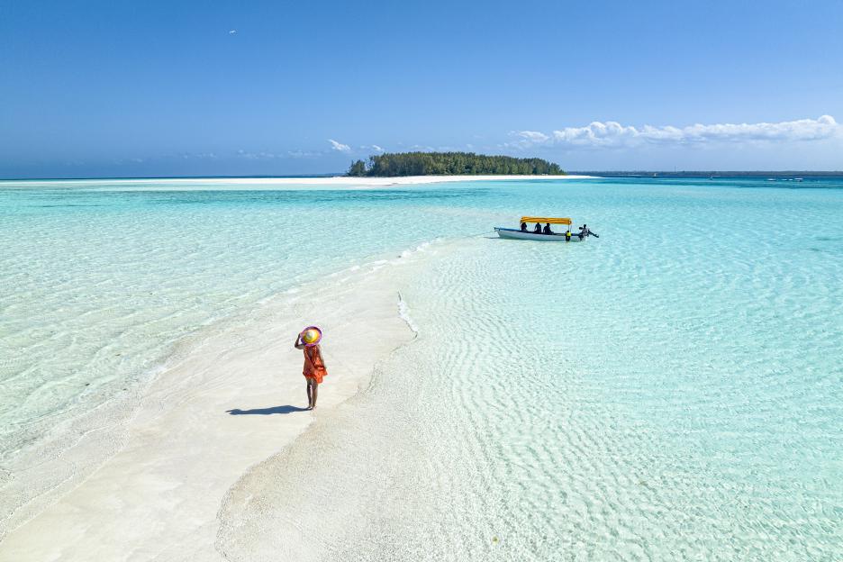 Aerial view of a woman in an orange dress and straw hat walking on a white sand bar in turquoise water, with a boat and distant island.