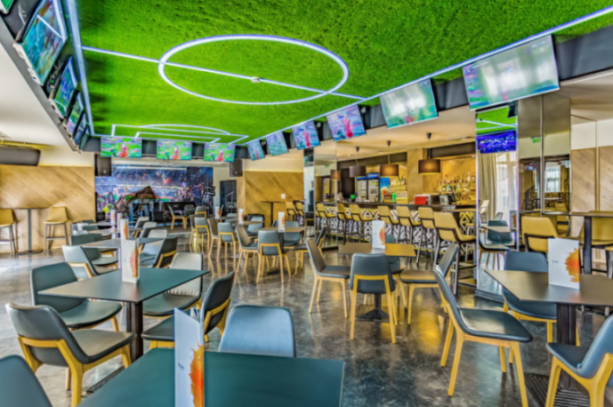 Indoor sports bar with green turf ceiling, multiple TV screens, tables, chairs, and a full bar.