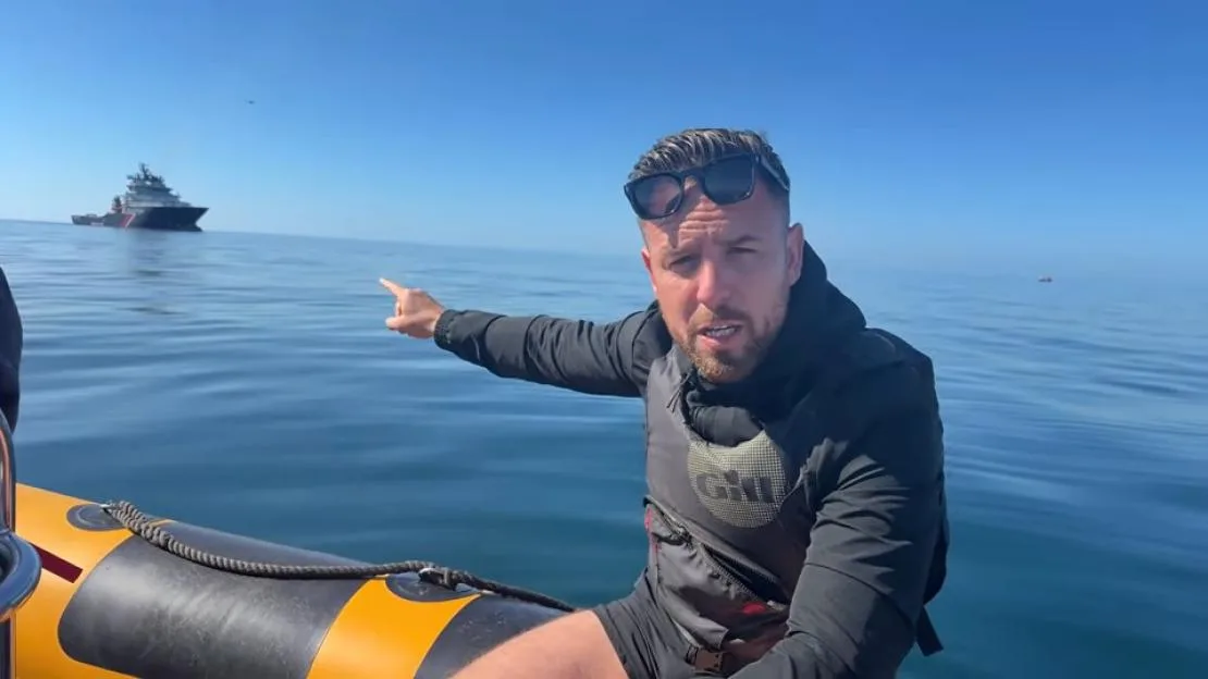 A man in a boat points towards a distant ship in the sea.